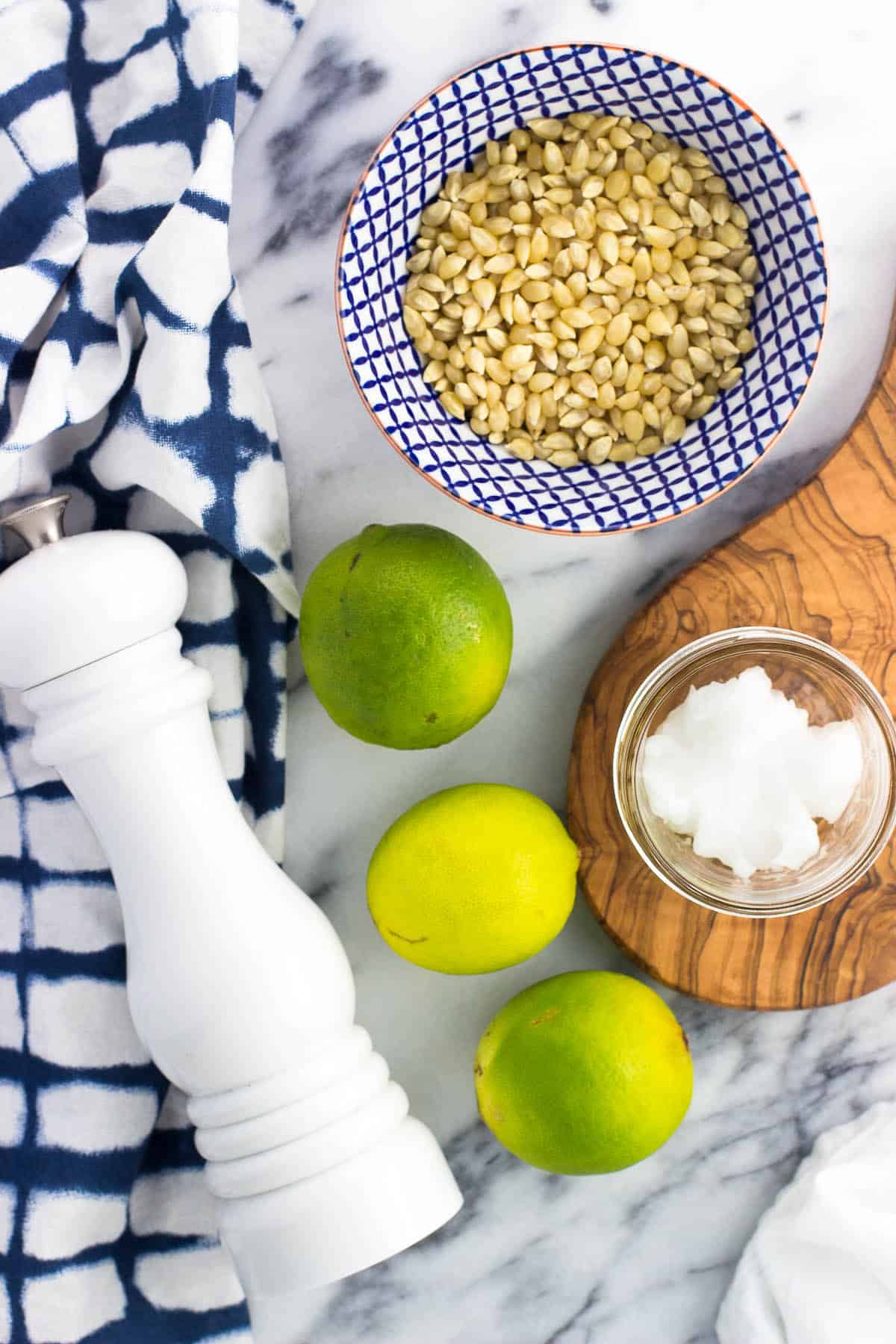 Three limes, a salt mill, a bowl of popcorn kernels, and a small glass jar of coconut oil on a marble board