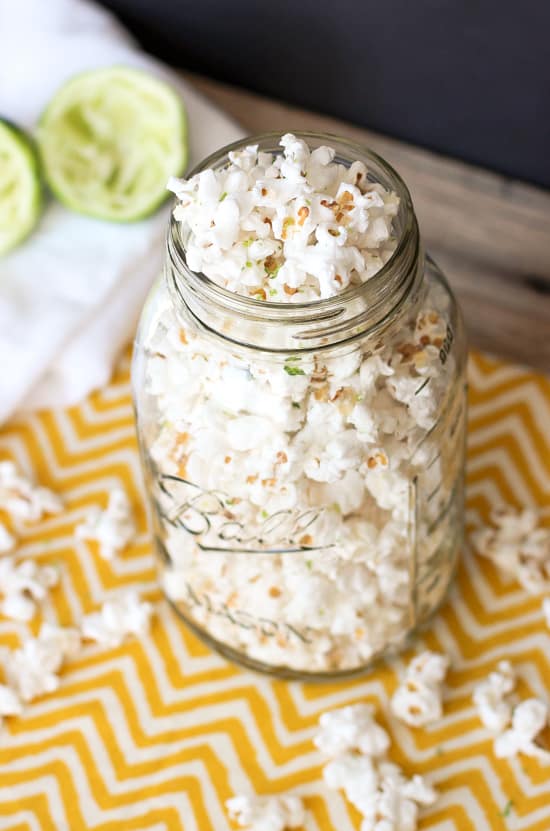 A large glass mason jar filled with popcorn on a dishtowel surrounded by loose popcorn and juiced lime halves