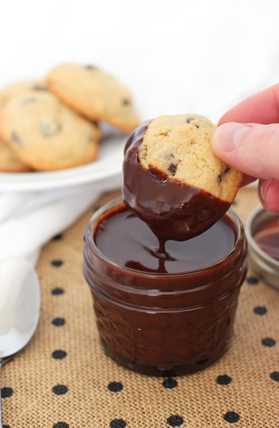 A hand dipping a chocolate chip cookie into a small jar of syrup.