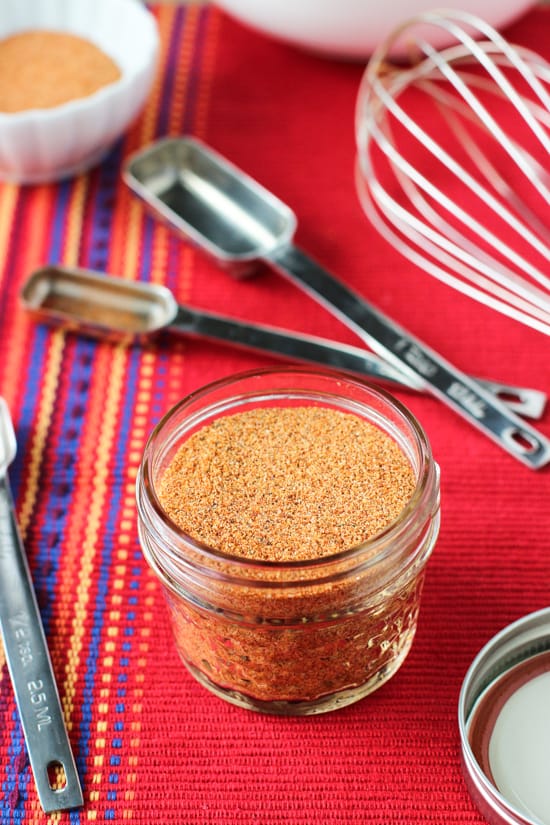 A small glass jar filled with the buffalo seasoning on a cloth placemat, surrounded by a whisk and measuring spoons