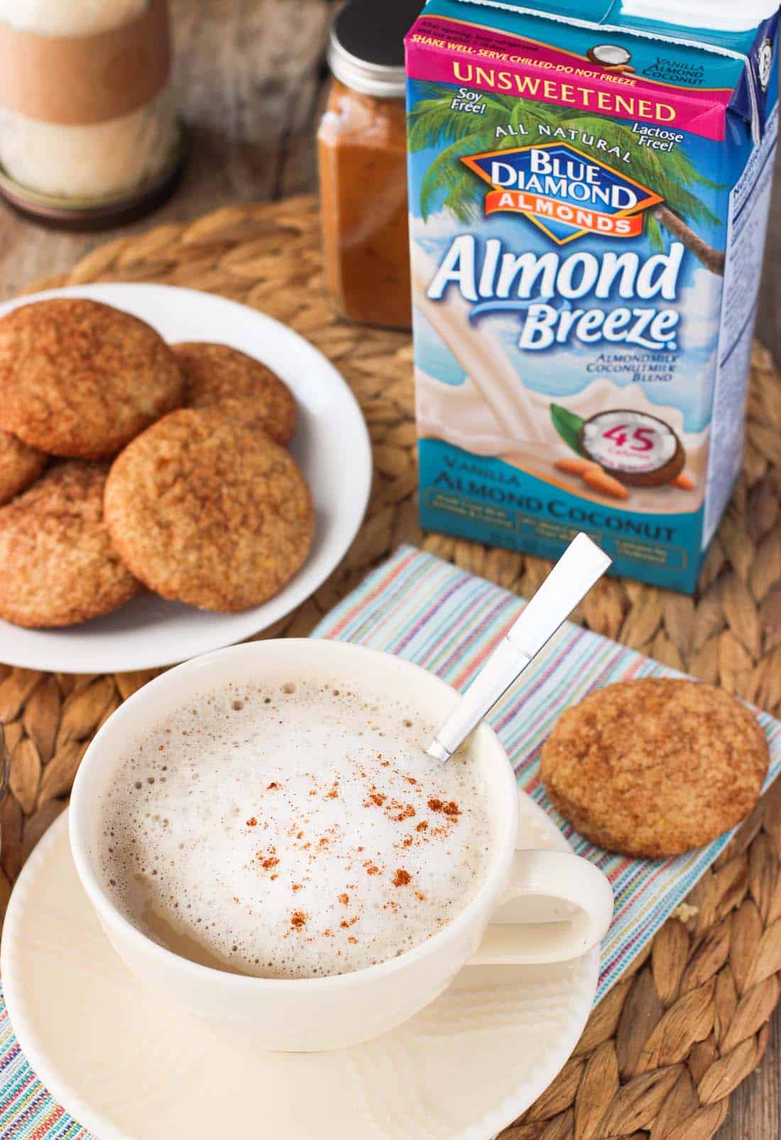 A latte in a mug with foamed milk and cinnamon served next to a plate of cookies with the Almondbreeze container in the background