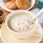 A latte in a wide mug with foamed milk and cinnamon to garnish. In the background is a plate of snickerdoodle cookies