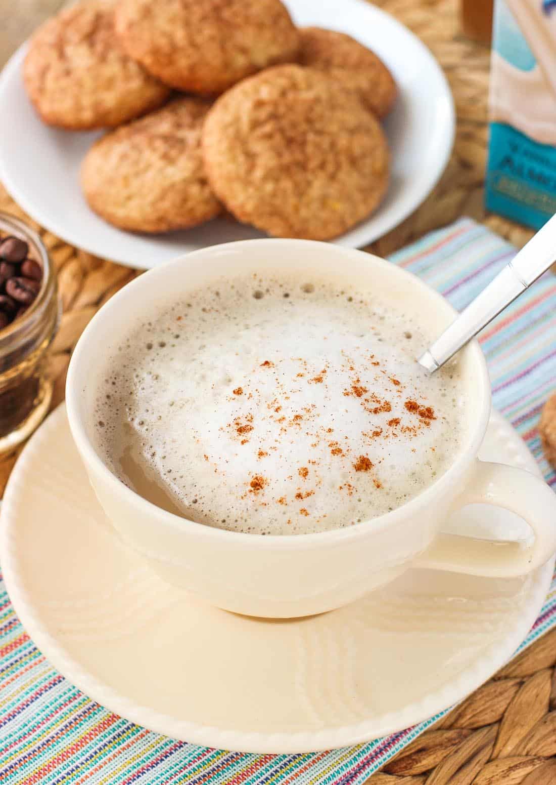 A latte in a wide mug with foamed milk and cinnamon to garnish. In the background is a plate of snickerdoodle cookies