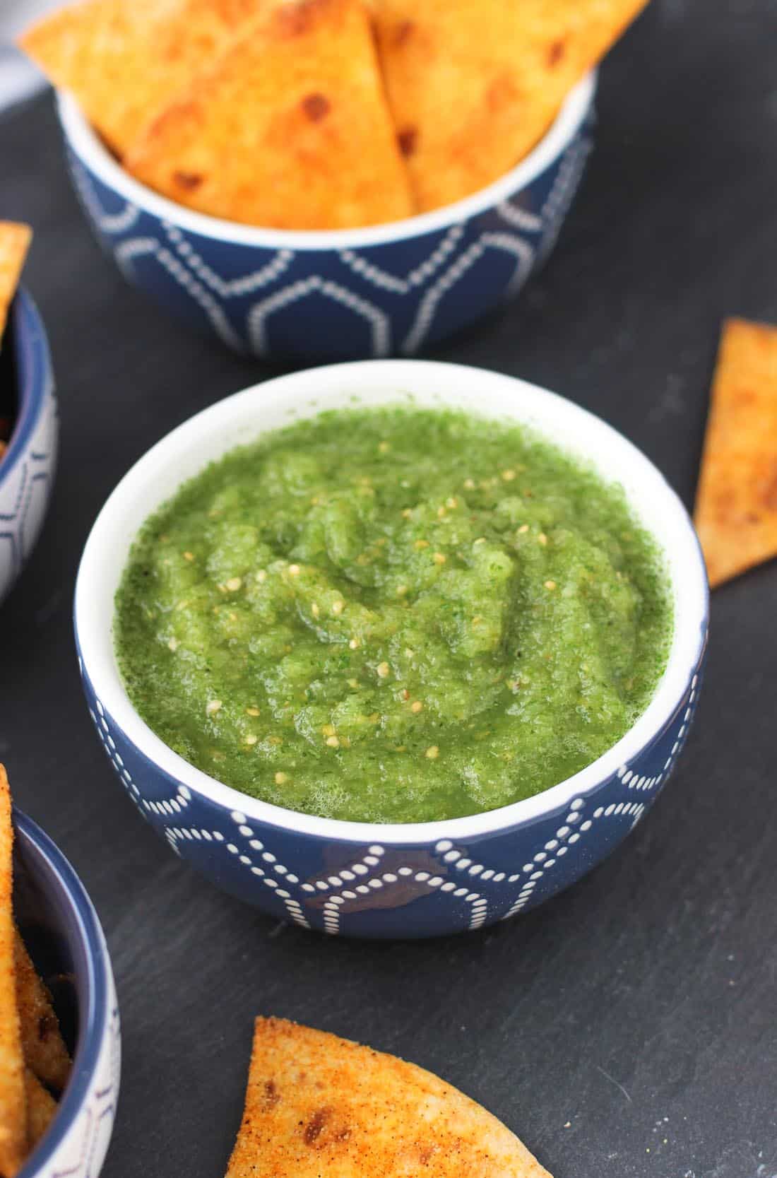 A bowl of salsa verde in front of bowls of tortilla chips.