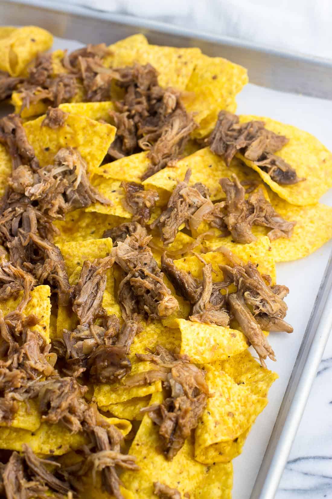 Tortilla chips laid out on a sheet pan topped with cooked pulled pork.