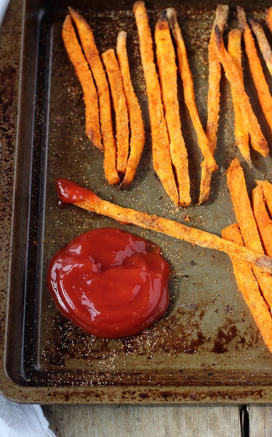 Sweet potato fries on a baking sheet with one dipped in a pool of ketchup.