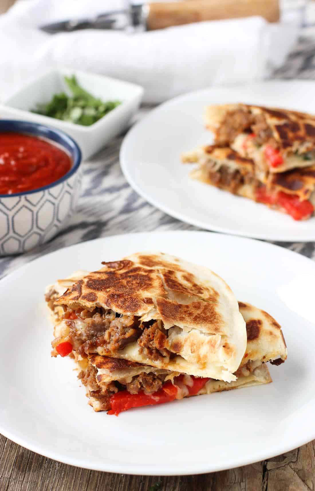 Stacked Italian sausage quesadillas on two plates with a dipping bowl of marinara sauce.