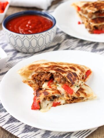 Stacked Italian sausage quesadillas on two plates with a dipping bowl of marinara sauce.