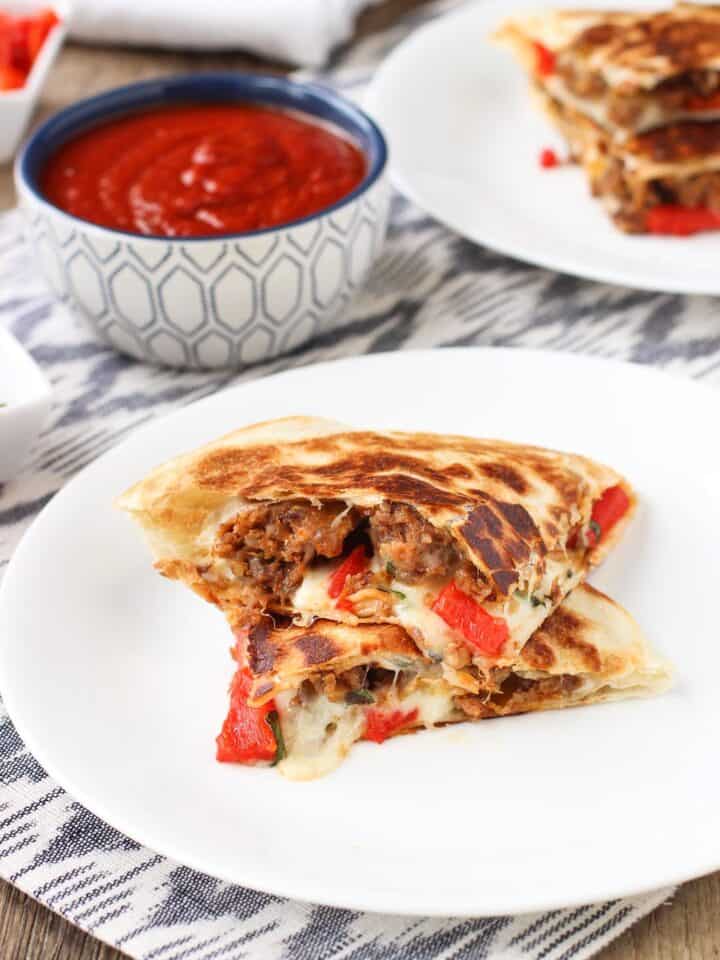 Stacked Italian sausage quesadillas on two plates with a dipping bowl of marinara sauce.