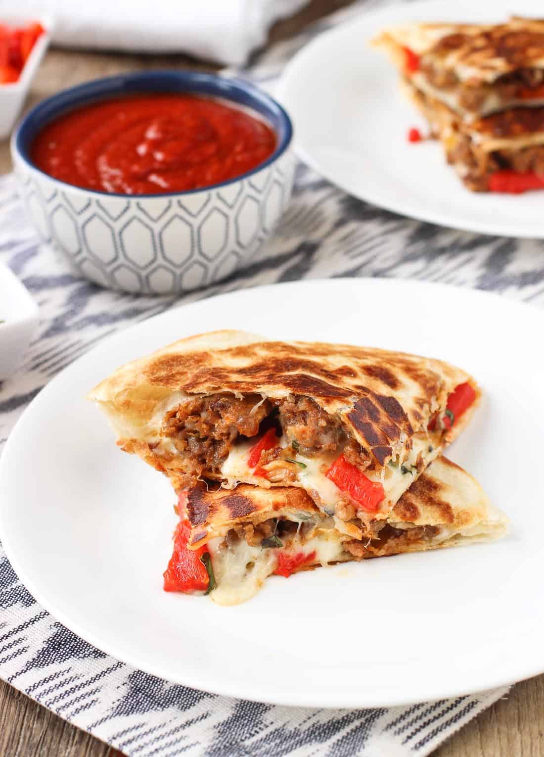 Stacked Italian sausage quesadillas on two plates with a dipping bowl of marinara sauce.