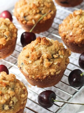 Muffins lined up on a metal cooling rack surrounded by fresh cherries with stems