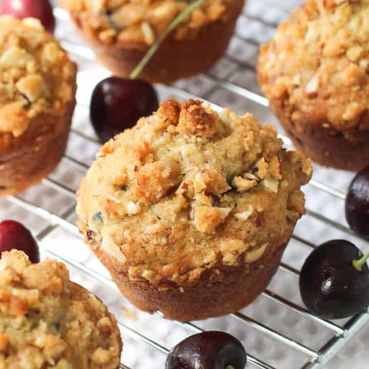 Muffins lined up on a metal cooling rack surrounded by fresh cherries with stems