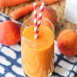 A smoothie in a tall glass with two straws sticking out with fresh peaches and whole carrots in the background