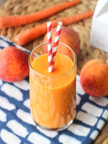 A smoothie in a tall glass with two straws sticking out with fresh peaches and whole carrots in the background