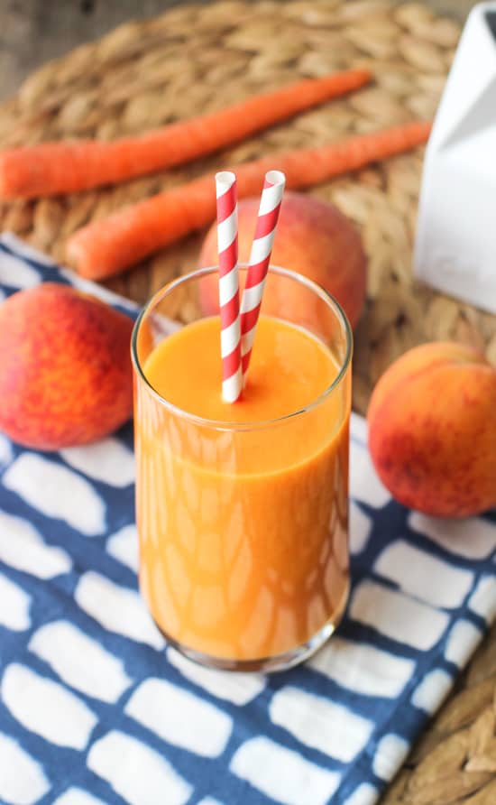 A smoothie in a tall glass with two straws sticking out with fresh peaches and whole carrots in the background.