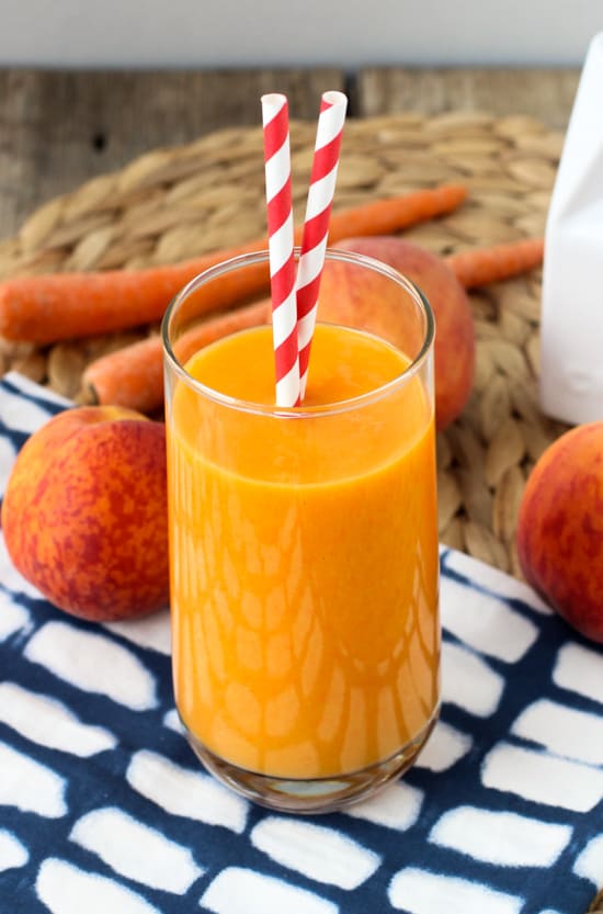 A peach mango smoothie in a tall glass surrounded by peaches and carrots.