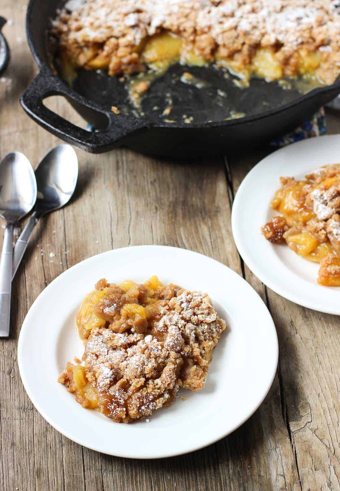 Two plates of portioned out peach crisp on a wooden table next to two dessert spoons and a cast iron pan