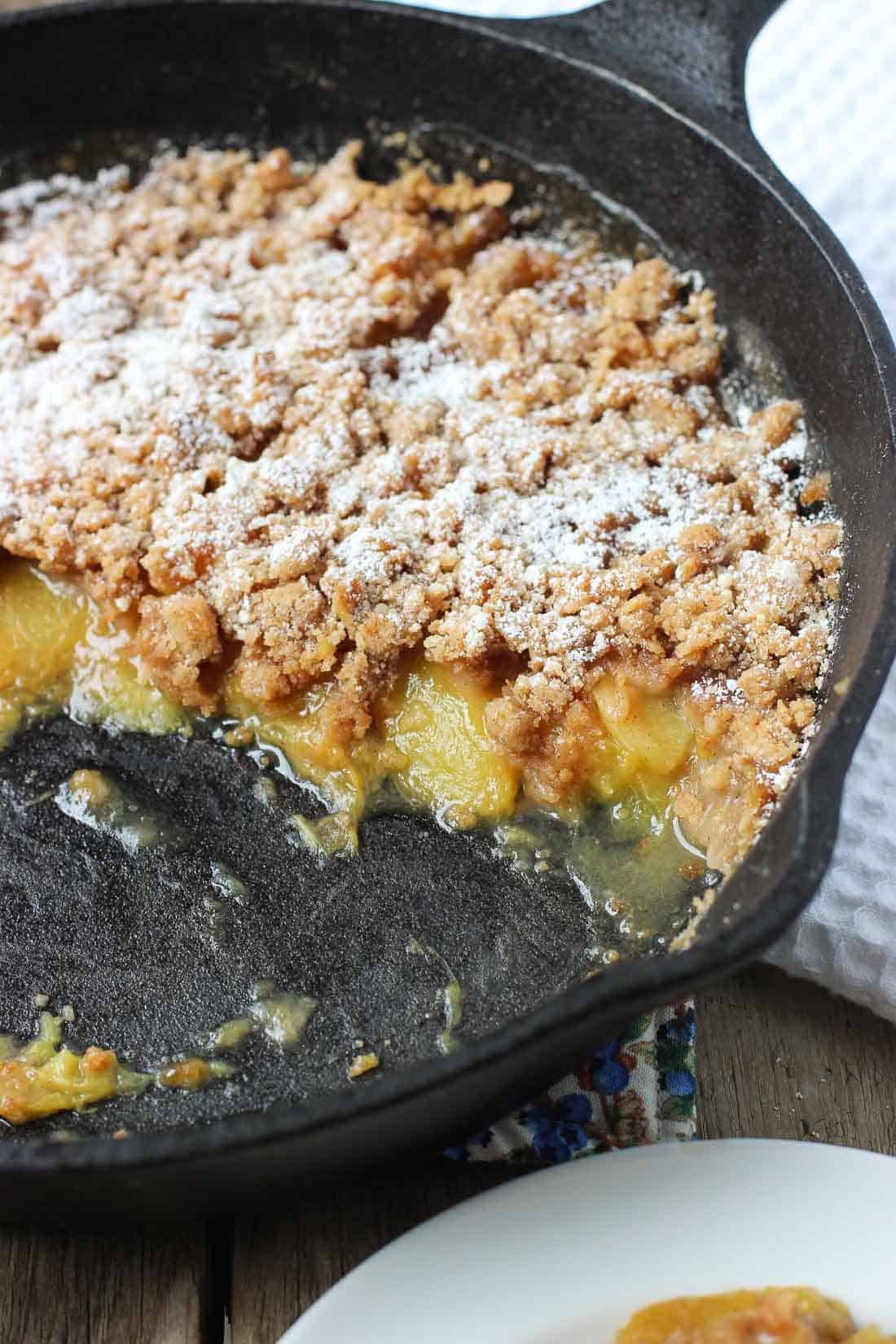 Half of a peach crisp in a cast iron skillet showing the peaches covered with a thick crumb layer dusted with powdered sugar