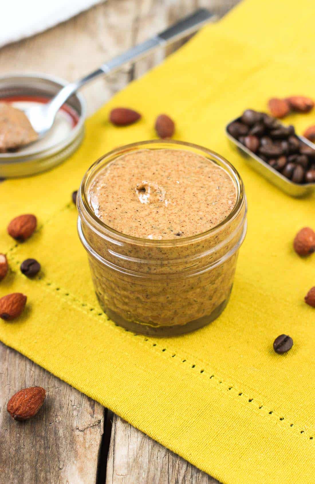 A small glass jar of almond butter on a dish towel surrounded by loose almonds and whole espresso beans