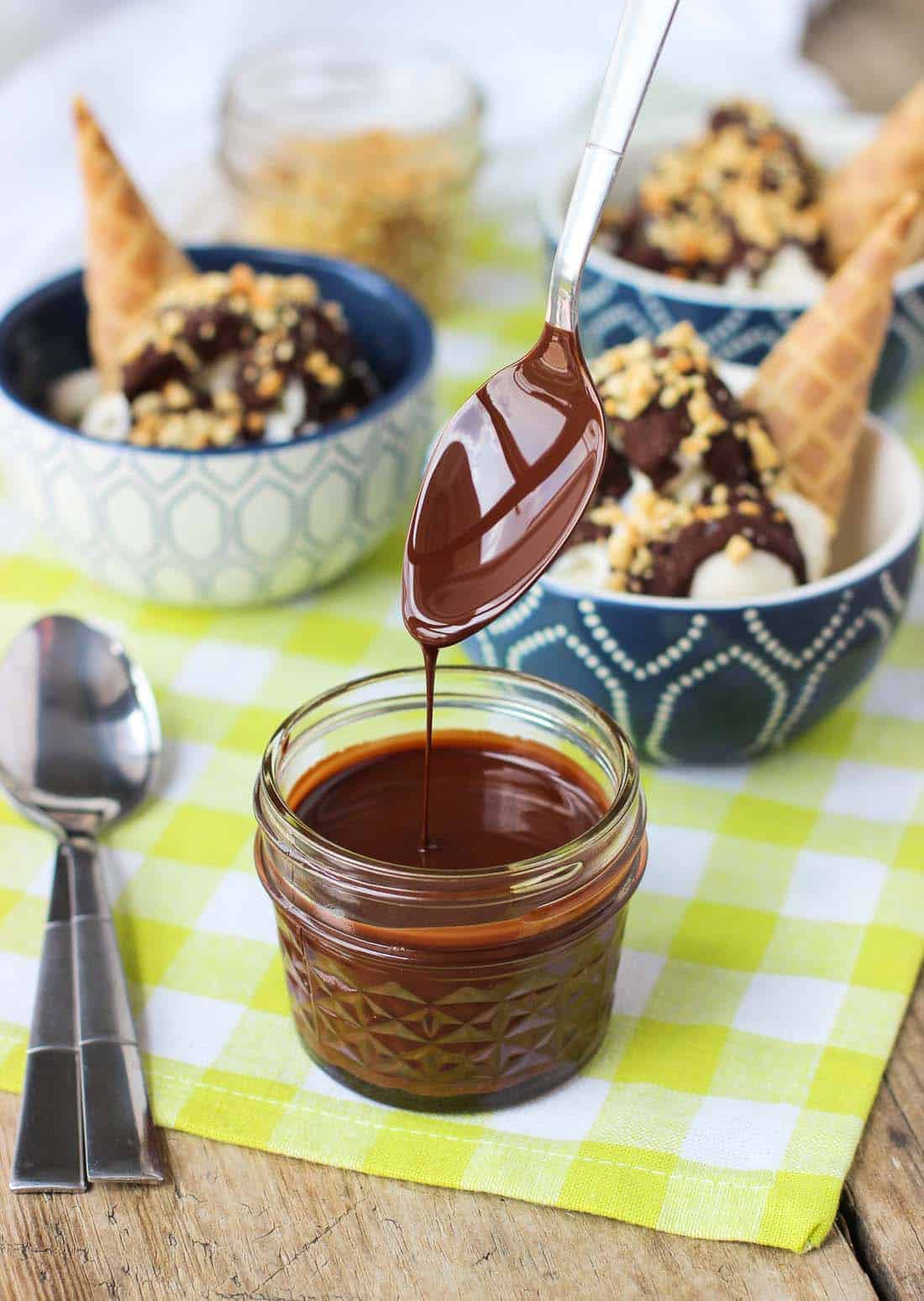 A spoon lifted out of a glass jar of chocolate magic shell, showing the thin mixture drizzling off the spoon