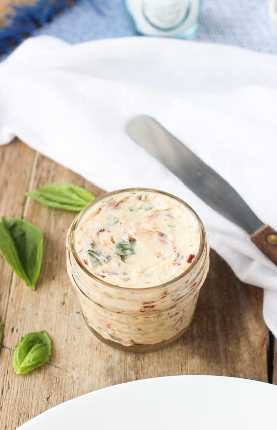 A small glass jar of compound butter next to a knife, a salt shaker, and a few scattered basil leaves