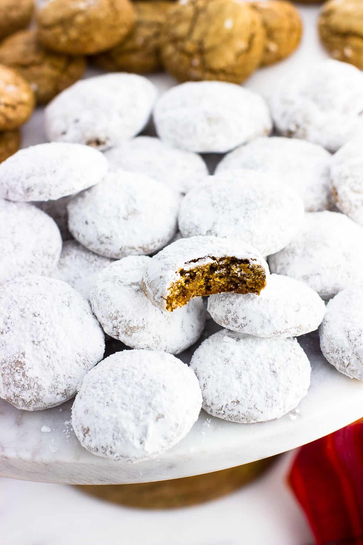 A batch of German spice cookies rolled in confectioners' sugar on a cake stand with a bite taken out of one.