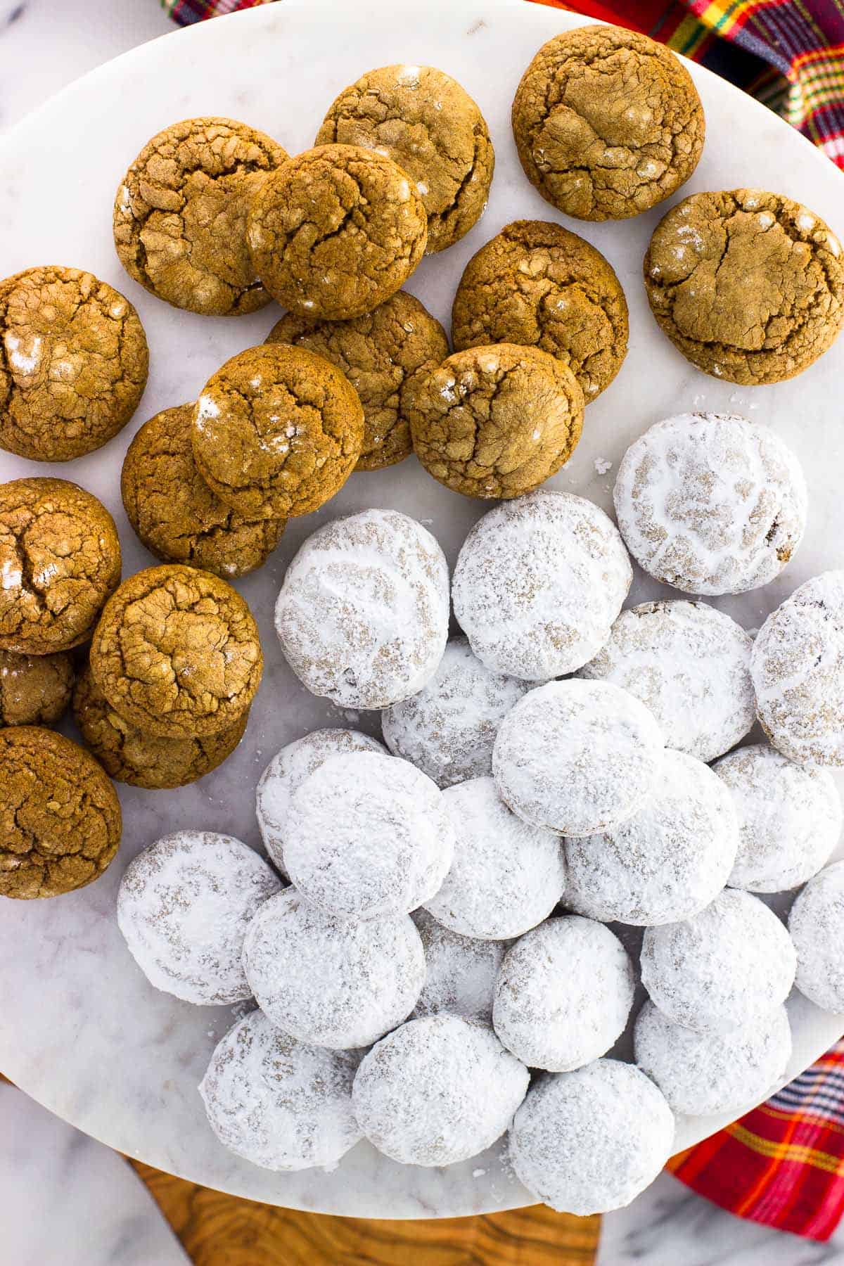 A batch of spice cookies on a cake stand, with half of the batch coated in confectioners' sugar and the other half not.