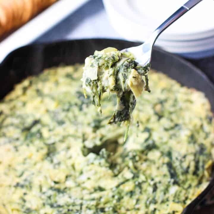 A spoon lifting a scoop of spinach artichoke dip out of the pan with crackers in the background.