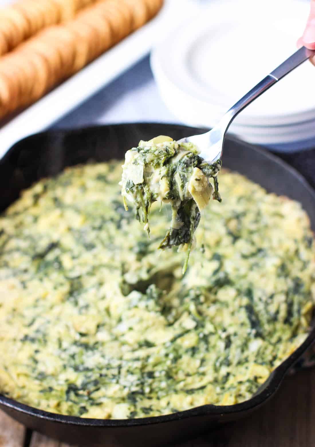 A spoon lifting a scoop of spinach artichoke dip out of the pan with crackers in the background.