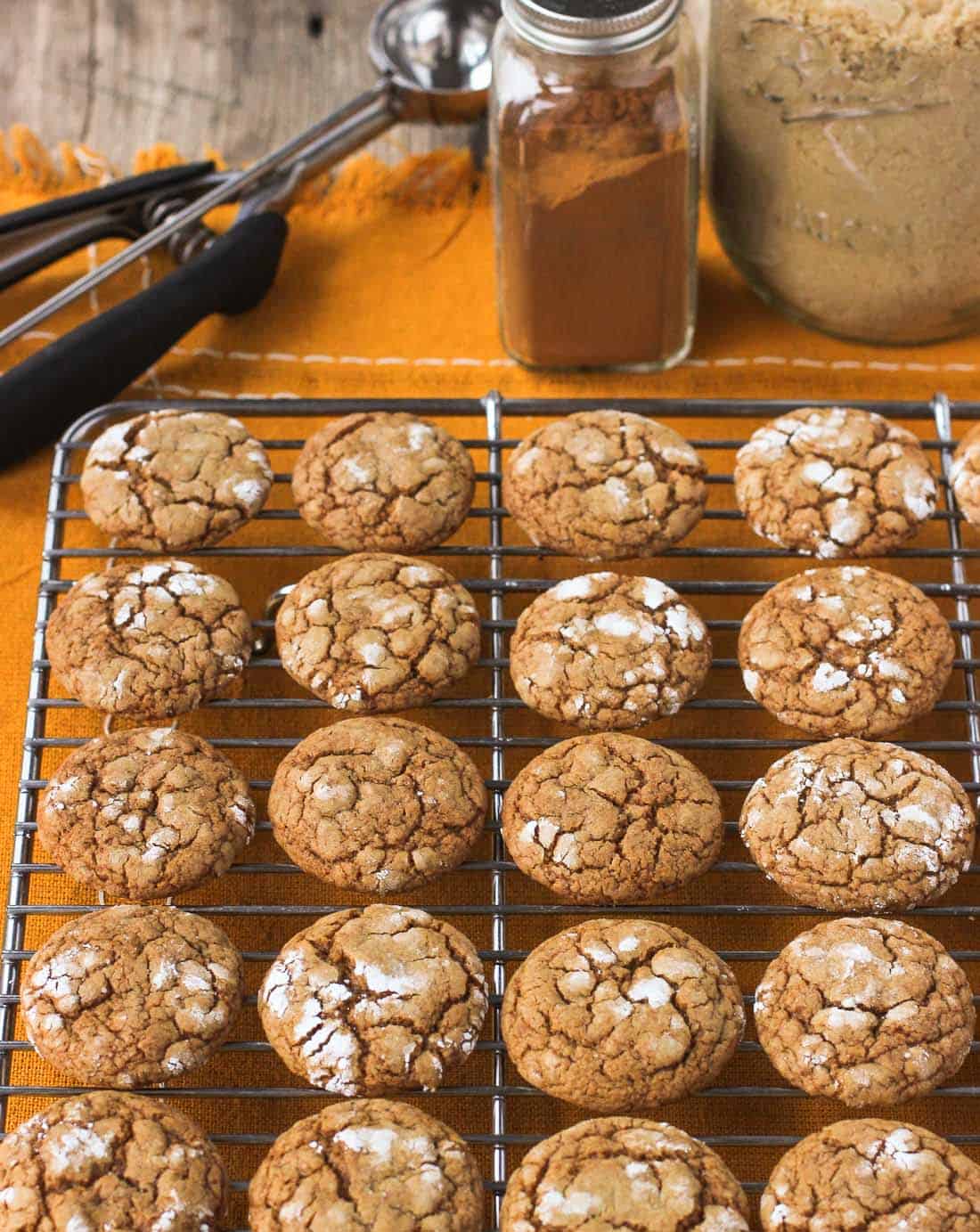 Lined up pfeffernüsse cookies on a wire rack next to a jar of cinnamon, a jar of brown sugar, and a cookie scoop.