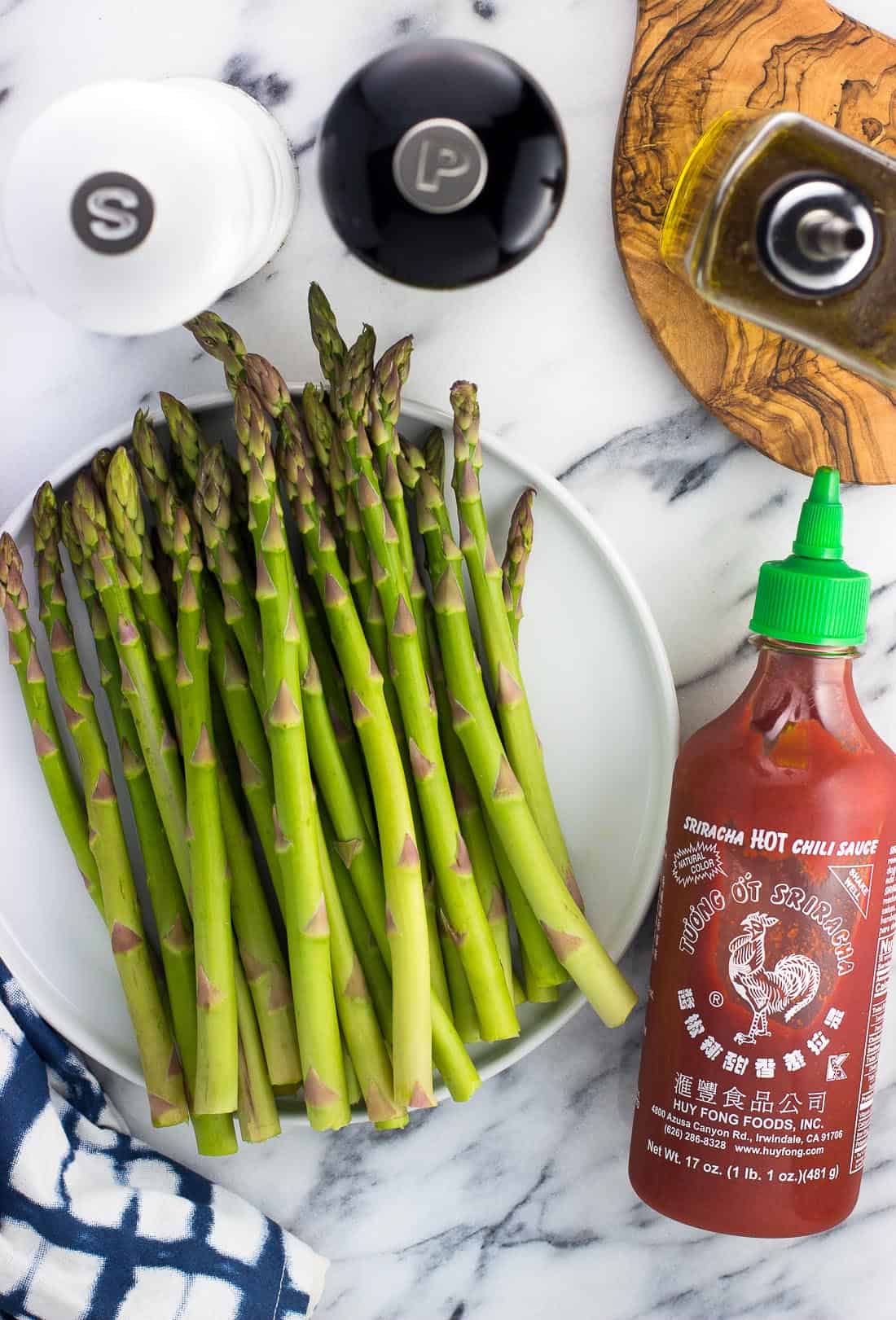 A bunch of raw asparagus, a bottle of sriracha, salt and pepper grinders, and a jar of olive oil on a marble board