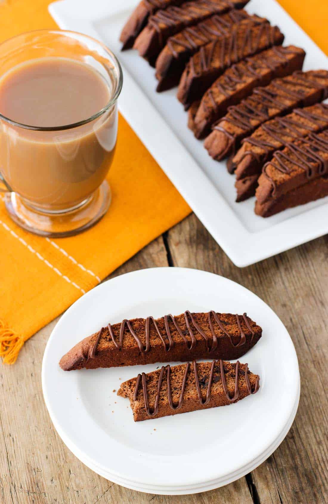 Two biscotti on a plate next to the tray of them and a mug of coffee.