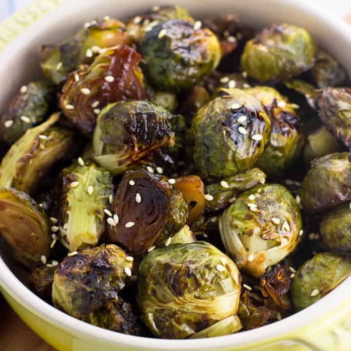 A small shallow dish filled with roasted brussels sprouts topped with sesame seeds in front of a dish towel