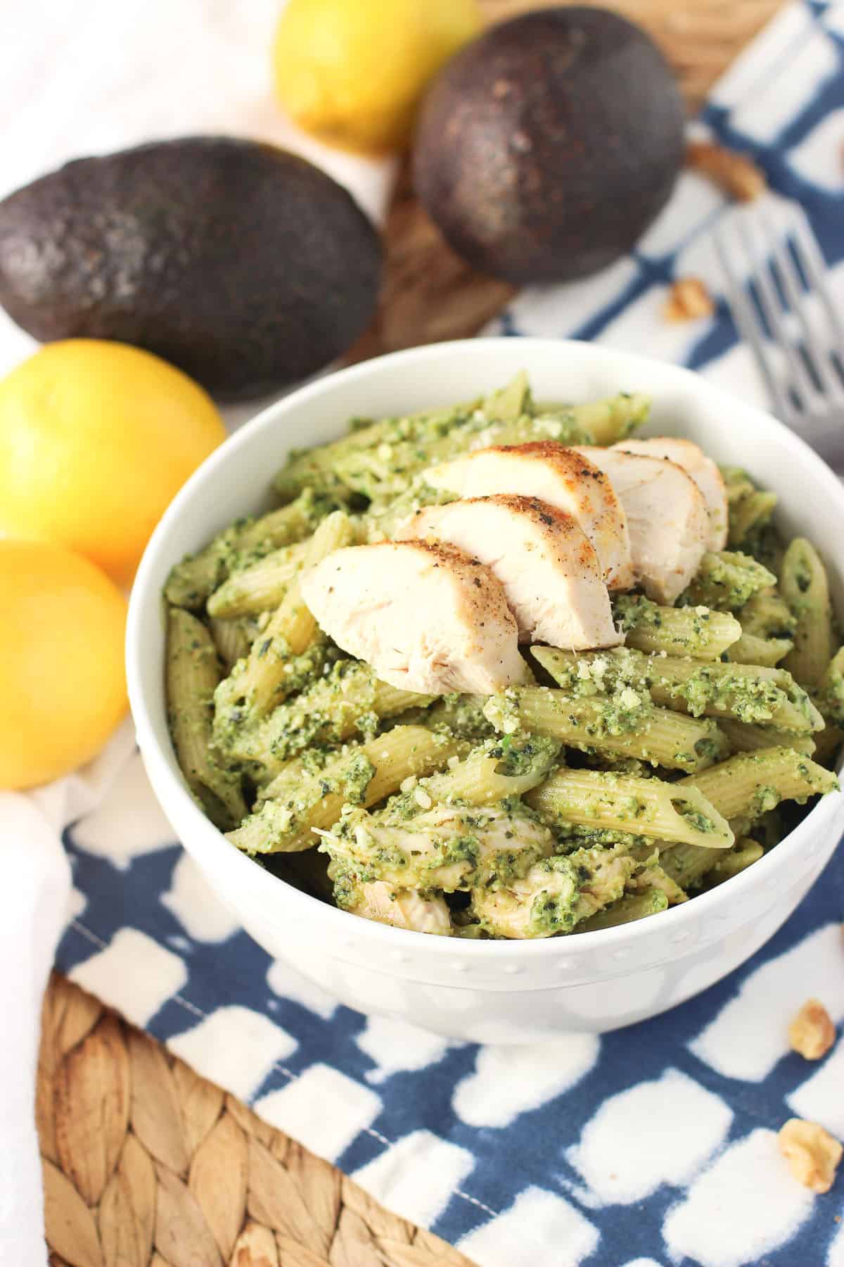 A bowl of pesto-covered penne with grilled chicken surrounded by lemons and avocados.