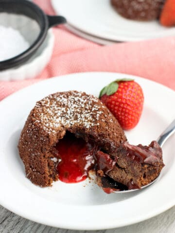 A lava cake on a dessert plate with a spoonful of cake removed to show the molten strawberry filling. The dish is garnished with a fresh strawberry.