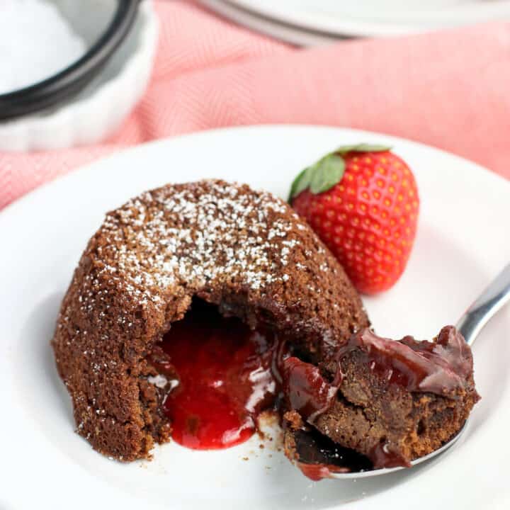 A lava cake on a dessert plate with a spoonful of cake removed to show the molten strawberry filling. The dish is garnished with a fresh strawberry.