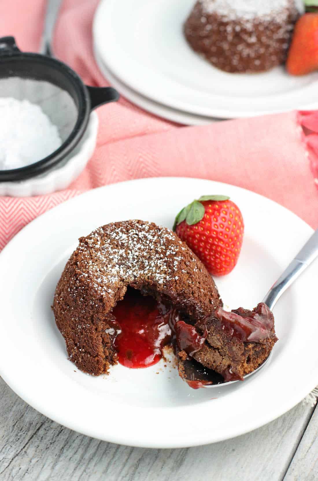 A lava cake on a dessert plate with a spoonful of cake removed to show the molten strawberry filling. The dish is garnished with a fresh strawberry.