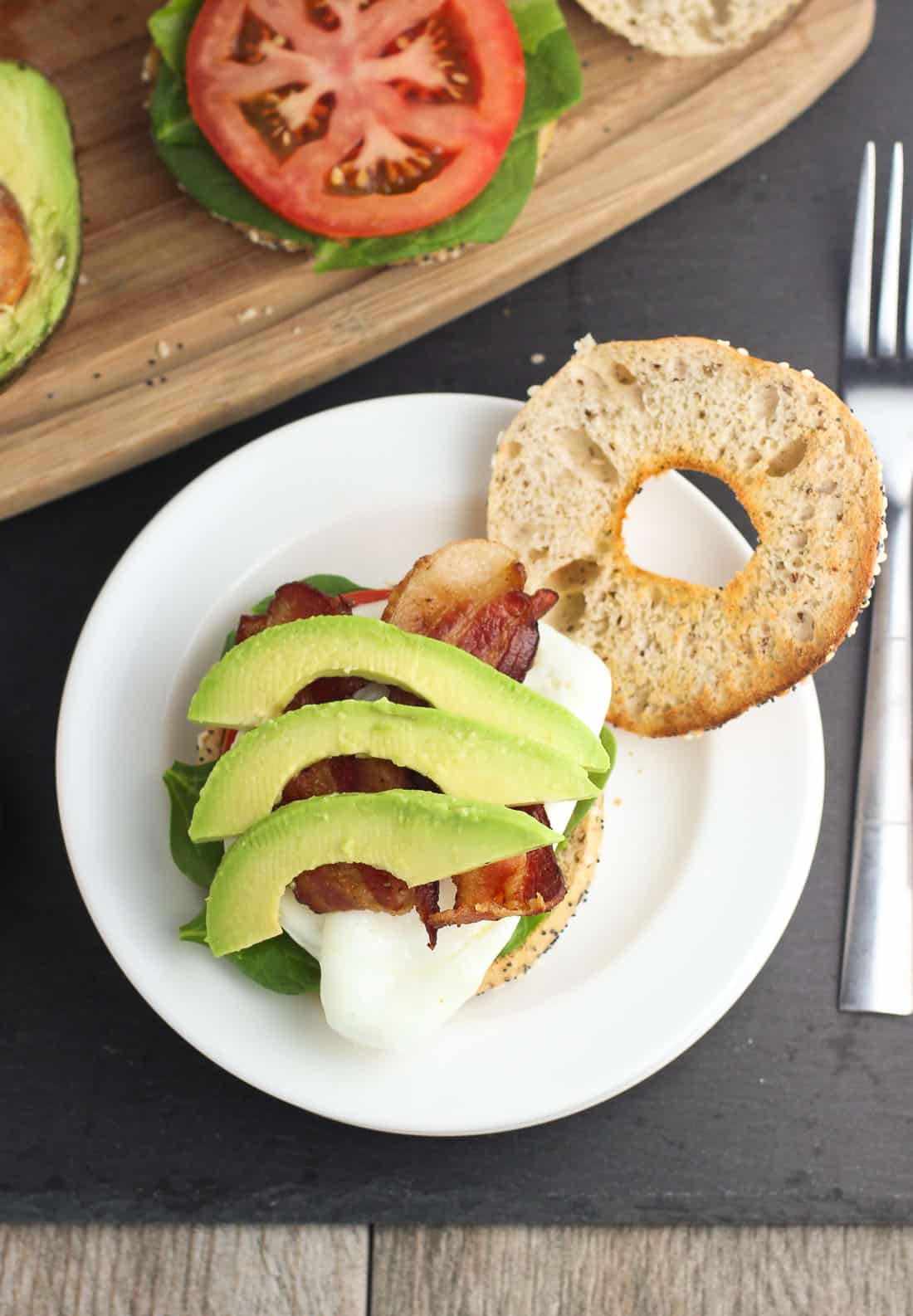 An open face breakfast sauce featuring lettuce, bacon, tomato slices, avocado slices, and an egg on a toasted bagel thin