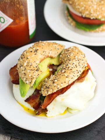 A BLT with avocado slices and egg on an everything bagel thin on a small plate next to a bottle of hot sauce