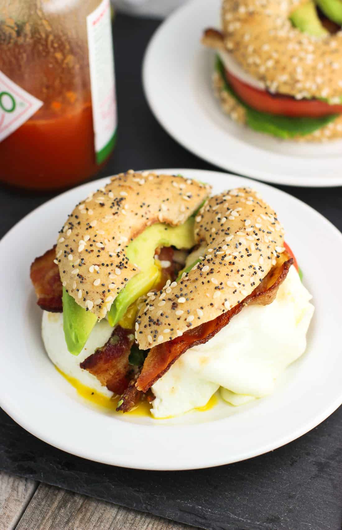 A BLT with avocado slices and egg on an everything bagel thin on a small plate next to a bottle of hot sauce