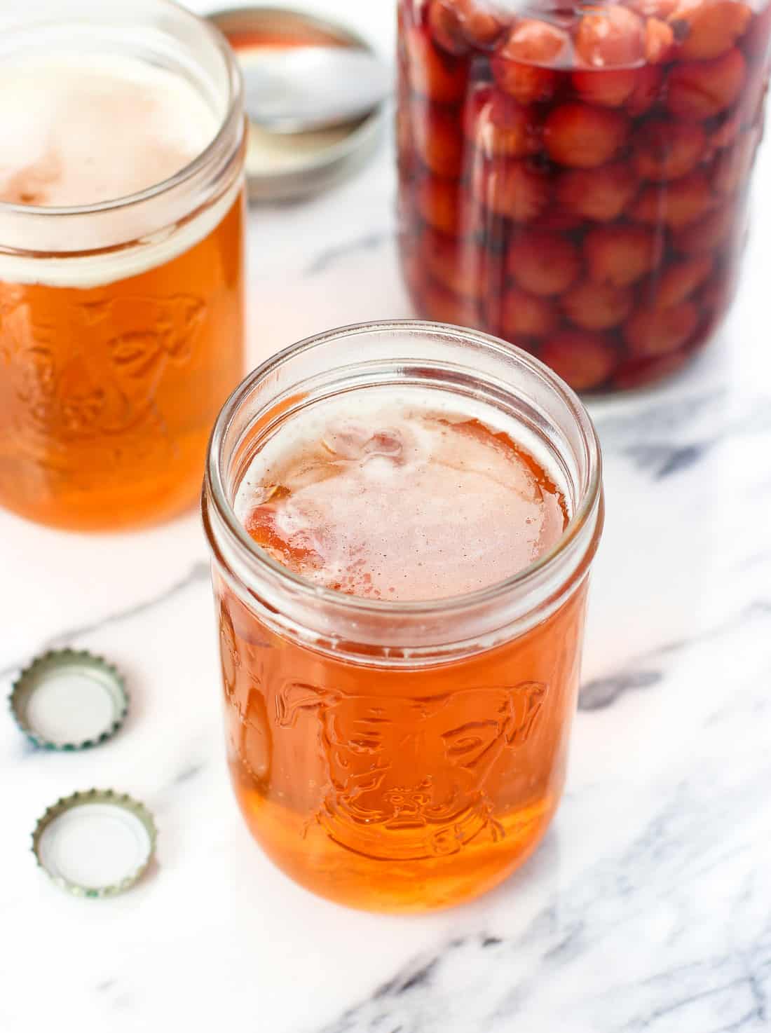 Two snakebites in beer glasses next to a large jar of maraschino cherries and two beer caps