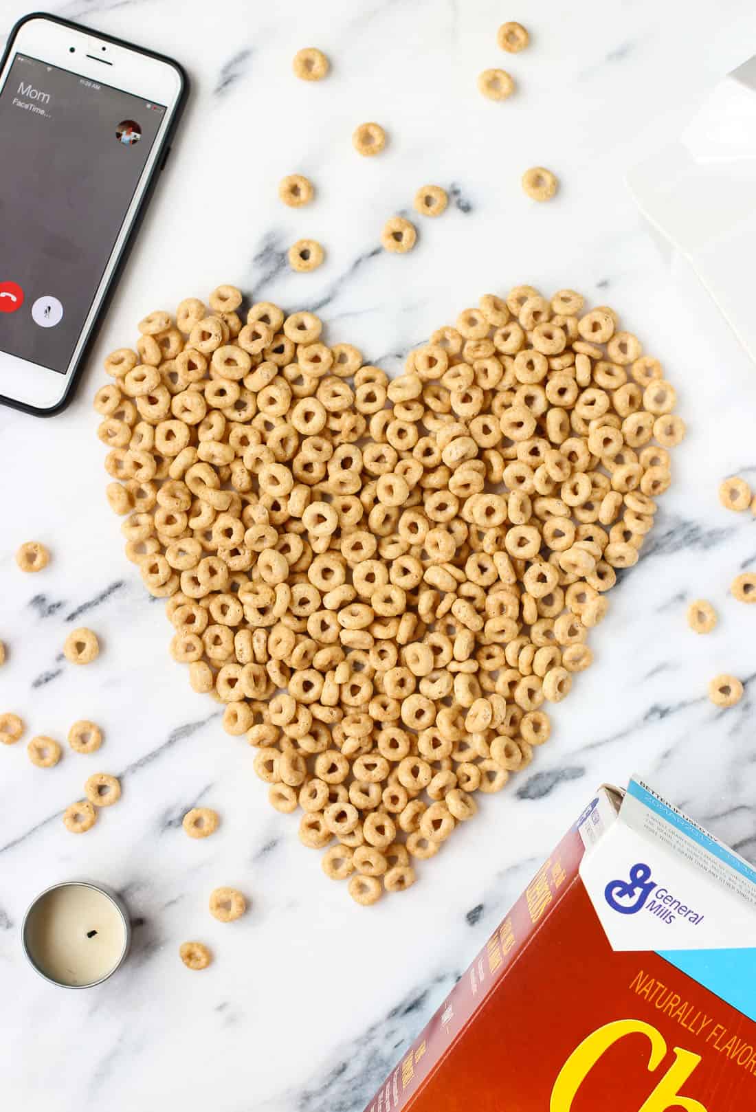 Cheerios arranged in a heart shape on a marble board surrounded by a phone, a candle, and the box.