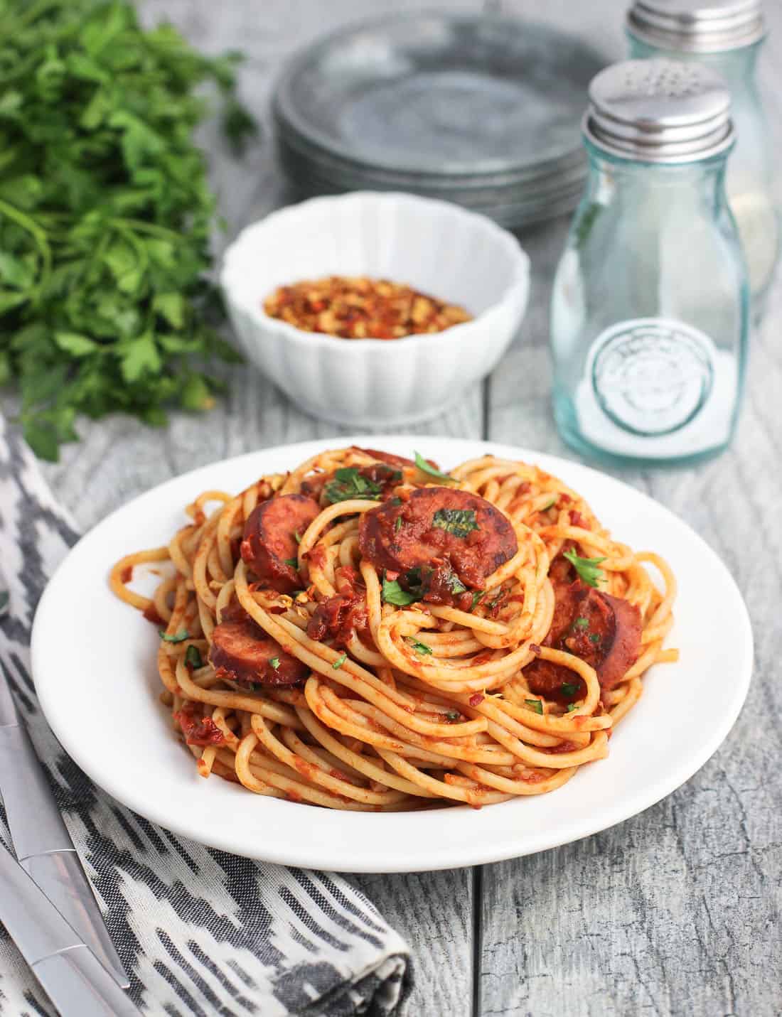 A serving of pasta on a dish surrounded by a salt shaker, red pepper flakes, and parsley.