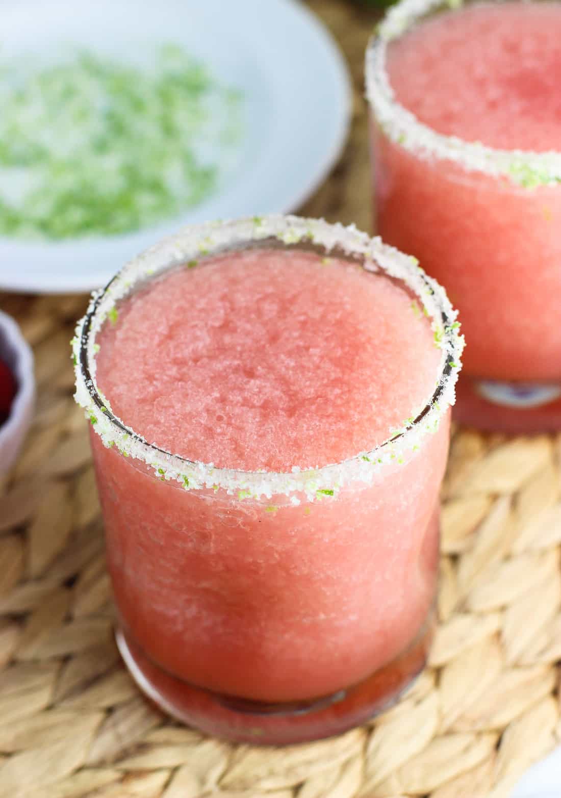 A close-up of frozen margarita with a salted lime rim with another margarita in the background along with the plate of salt and lime zest