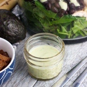 A small glass jar of dressing in front of a salad bowl and salad ingredients.