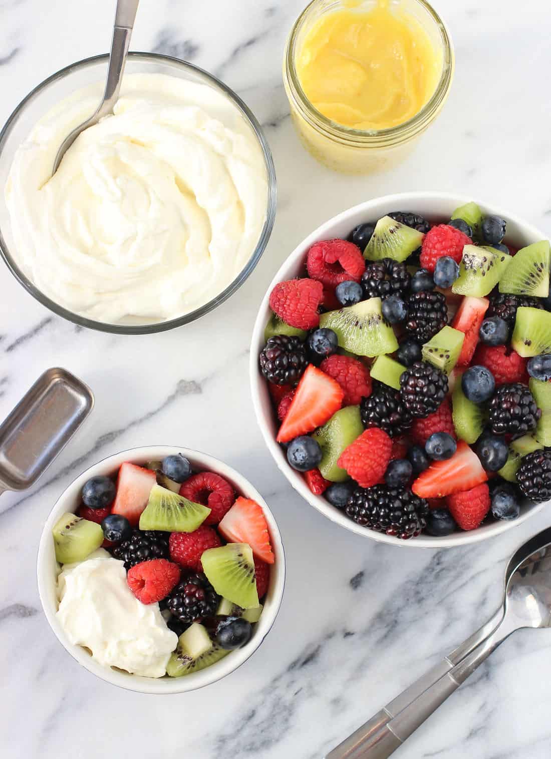 A bowl of whipped cream, a jar of curd, and two bowls of fruit salad on a marble board, with a dollop of whipped cream on the smaller bowl.