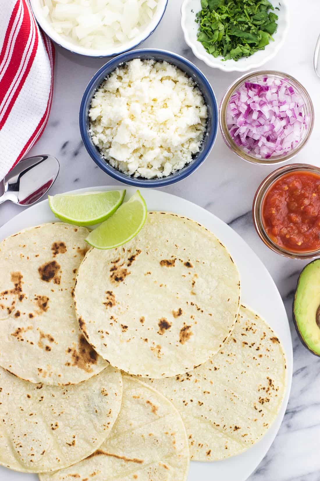 An overhead shot of all the taco fixings, including tortillas, cotija cheese, red onion, salsa, lime wedges, and avocado.