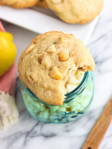 A white chocolate macadamia nut cookie with a bite taken out of it on top of a frosted glass jar of white chocolate chips