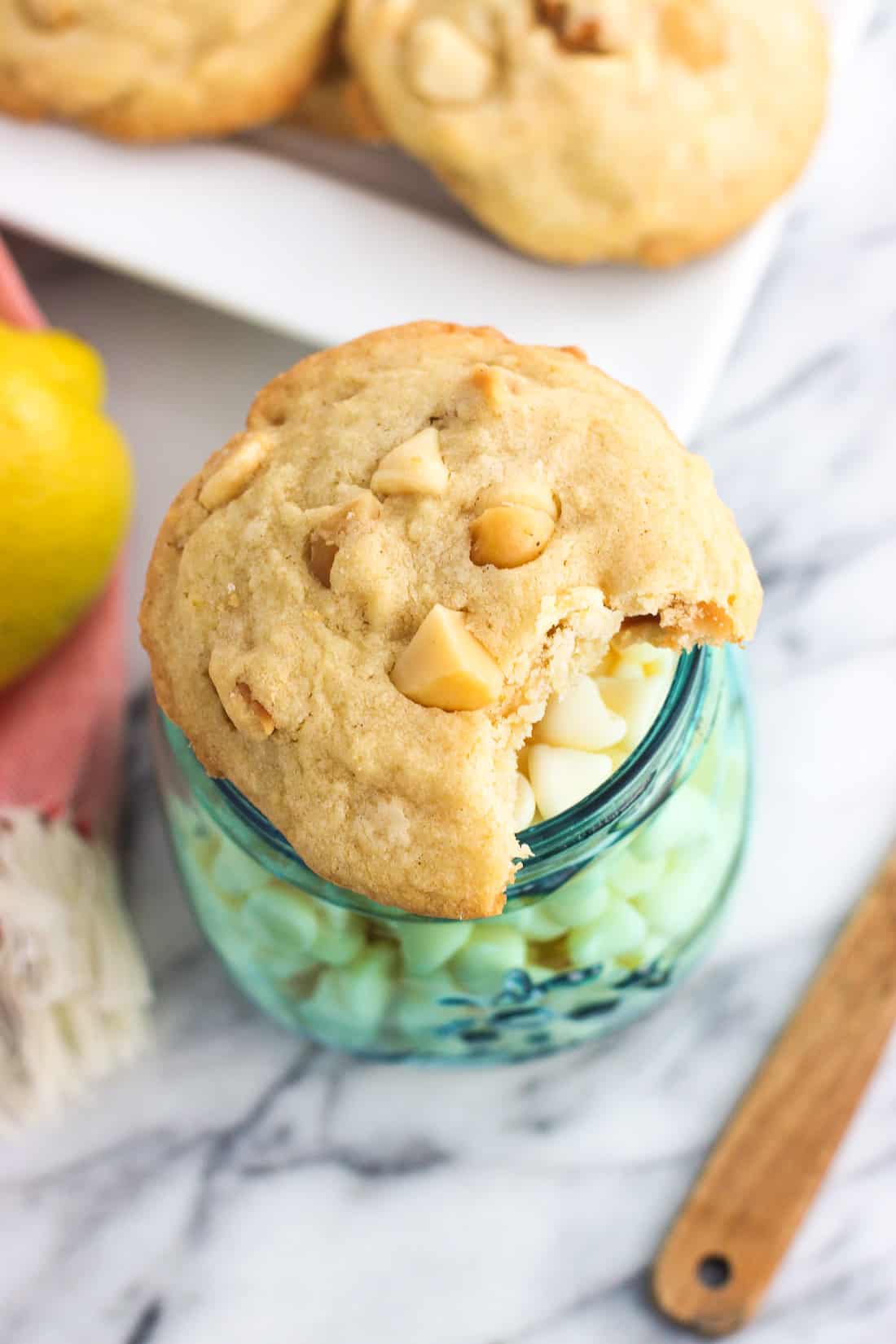A lemon white chocolate macadamia nut cookie with a bite taking out of it on top of a jar of white chocolate chips.