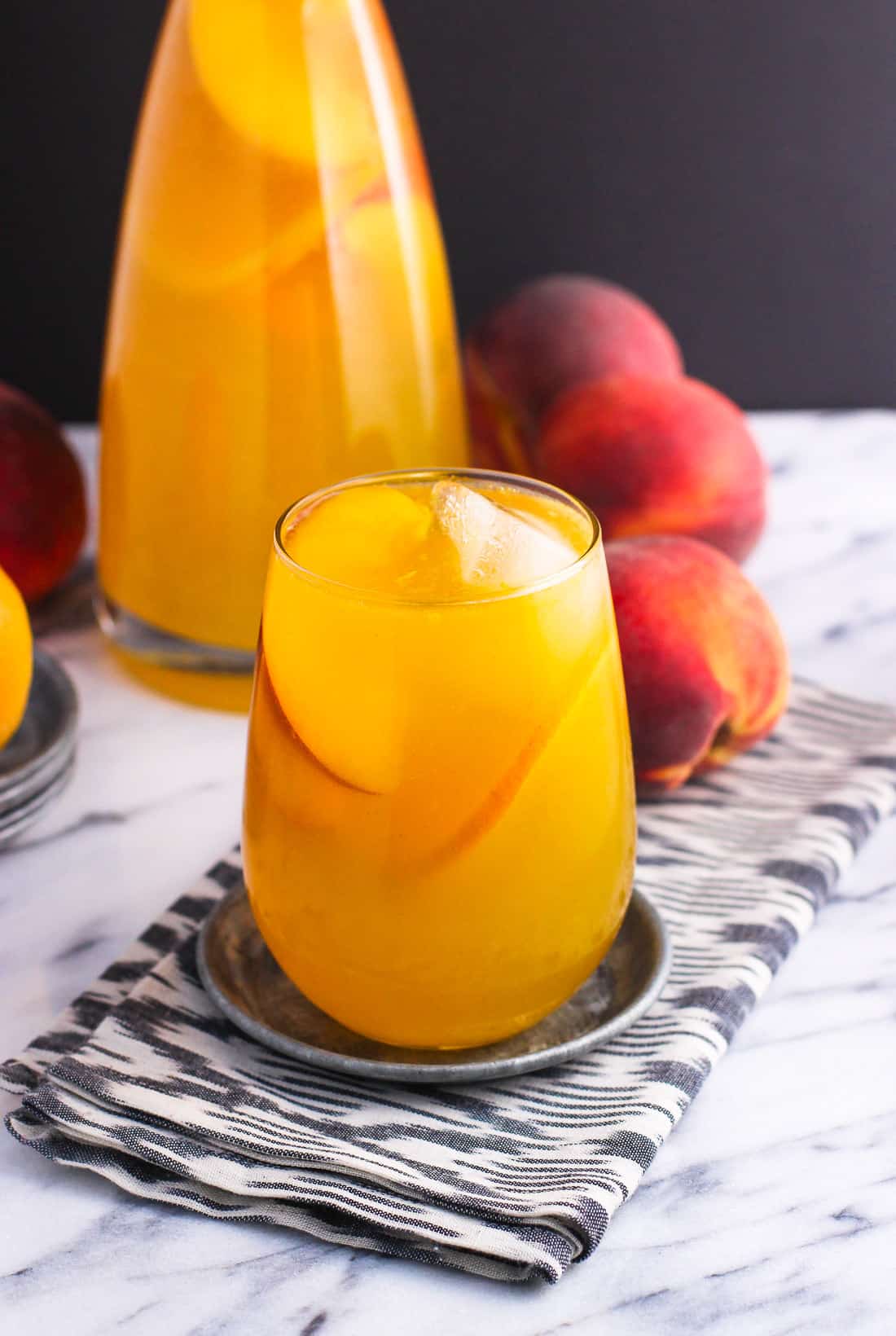 A glass of peach mango sangria garnished with ice cubes and peach slices in front of a carafe of sangria and peaches